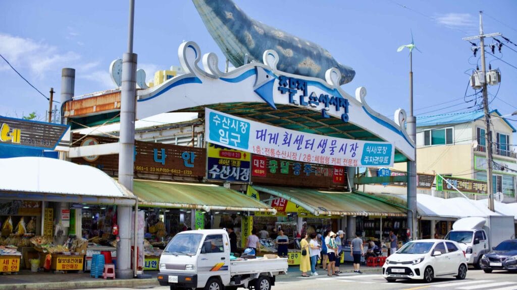 The entrance to Jumunjin Traditional Market, marked by a large whale sculpture atop the awning, welcomes visitors to a bustling scene of stalls selling an array of seafood products, including dried fish and squid.