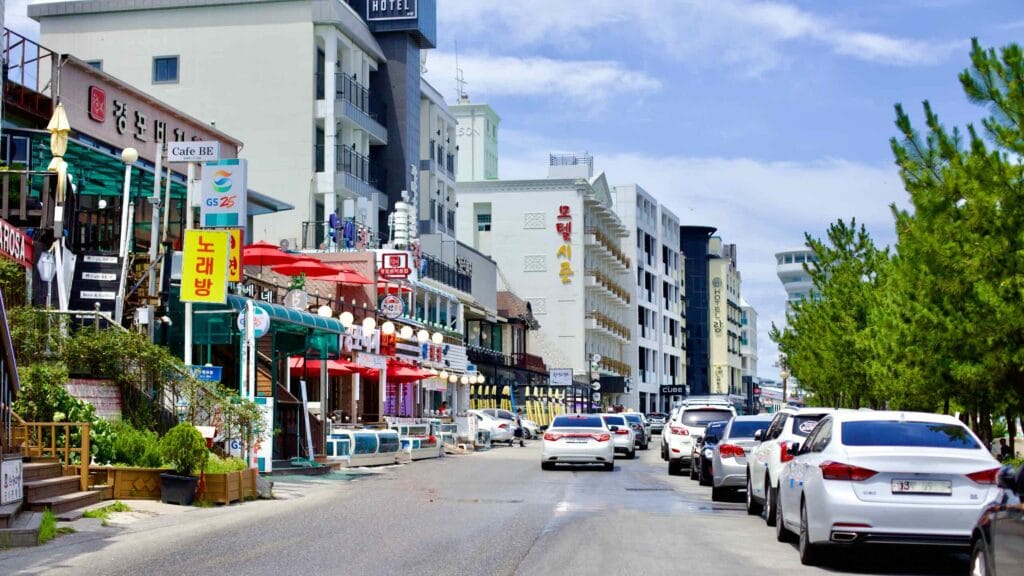 The bustling coastal street nestled between Gyeongpodae Lake and Gyeongpodae Beach, lined with seafood restaurants, convenience stores, and hotels.