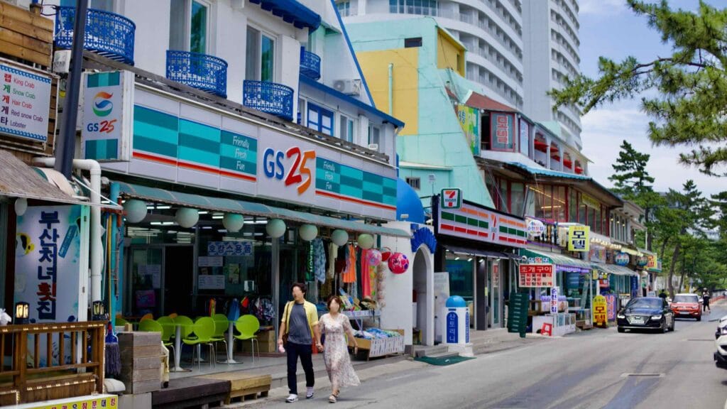 A lively street of convenience stores and restaurants fronts Gyeongpodae Beach, with the towering Skybay Hotel Gyeongpo set against a clear blue sky.