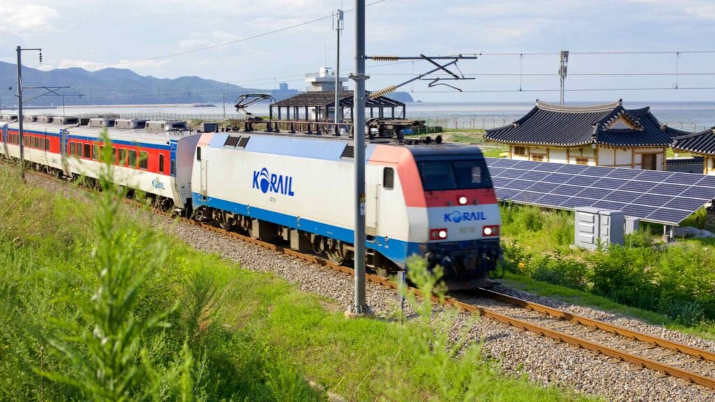 A Korail train passes by the edge of Mangsang Beach Hanok Village, with solar panels and the East Sea in the backdrop, blending tradition with modern sustainability.