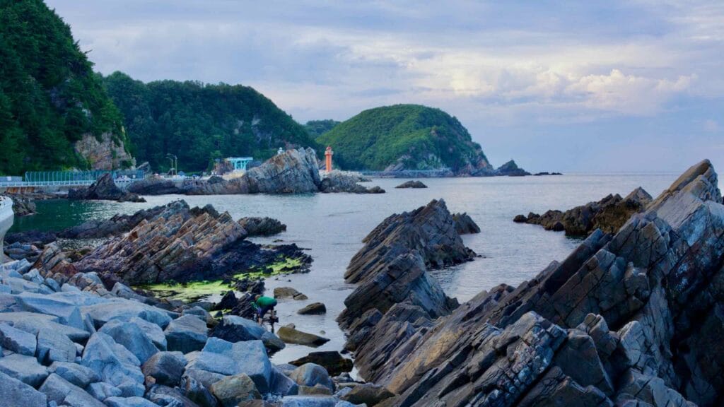 Angular rocks in the waters off of Heonhwa Road with the picturesque Simgok Port, a round green seaside hill, and a small red lighthouse on the breakwater in the background.