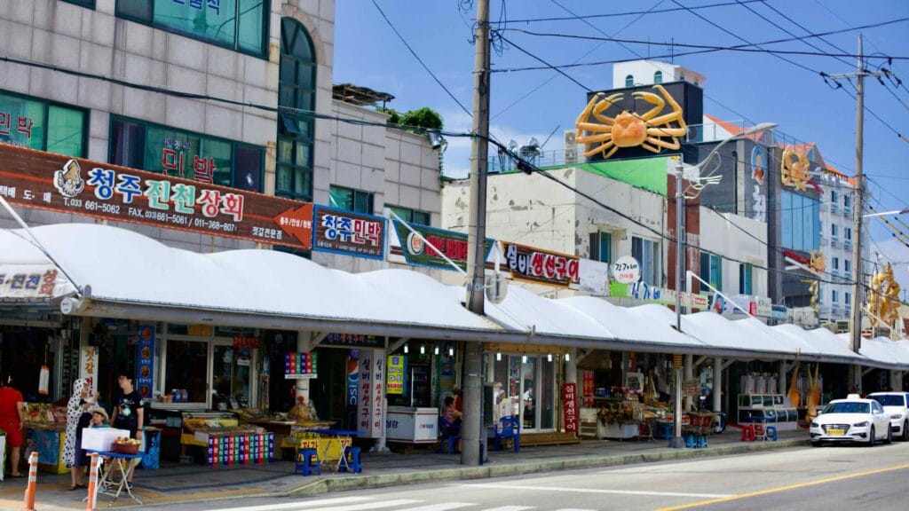 The bustling seaside market at Jumunjin Port, alive with seafood restaurants and storefronts, crowned by a large crab sculpture, inviting visitors to explore the flavors of the East Sea.