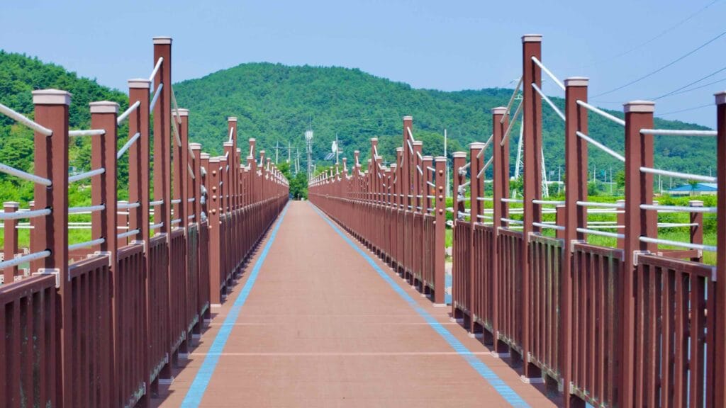 The top deck of the Bukcheon Railway Bridge boasts a high red fence and a rope running along its length, with blue lines marking a dedicated bike path.