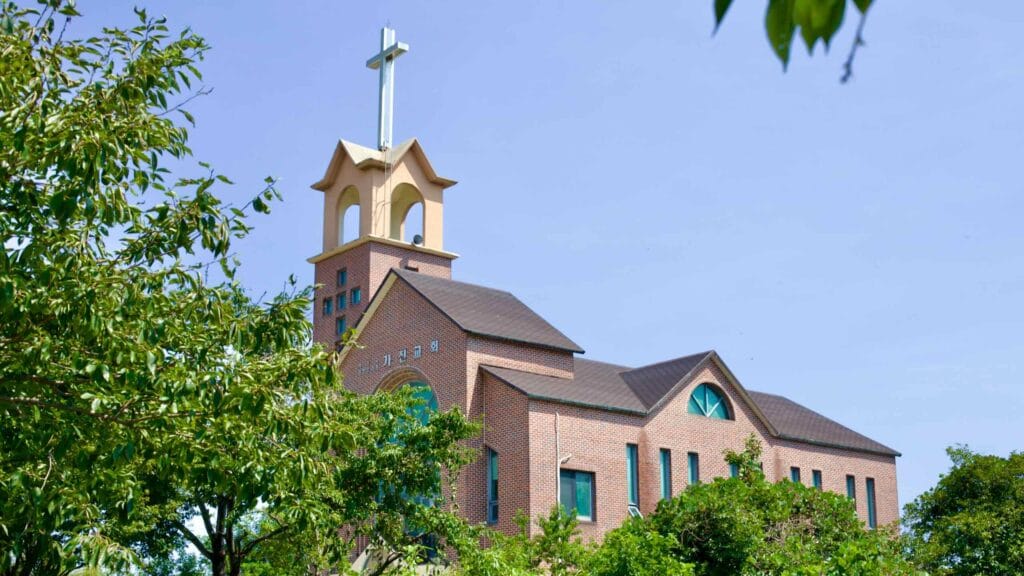 Gajin Church features a cross atop a three-story tall bell tower, with its main building nestled below, situated near Gajin Port under a clear blue sky.