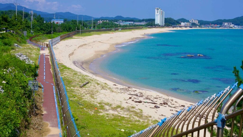 Geojin Village Beach unfolds with its sandy shores and blue waters, with a bike path bordered by a metal fence separating it from the beach.