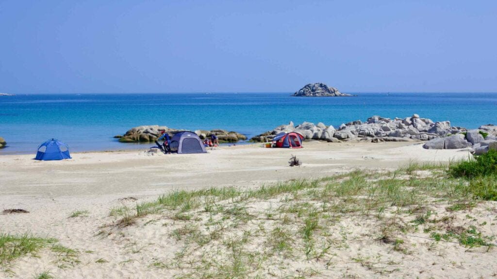 Sampo Beach presents a serene setting with tents pitched on its flat sandy shores, against the backdrop of Geomeundo Island, a small rocky island in the East Sea.