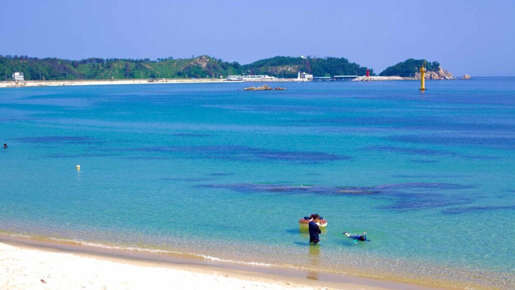 A family swims in the clear blue waters of Gonghyeonjin Beach, where white sands form a wide curving bay against a backdrop of lush hills.