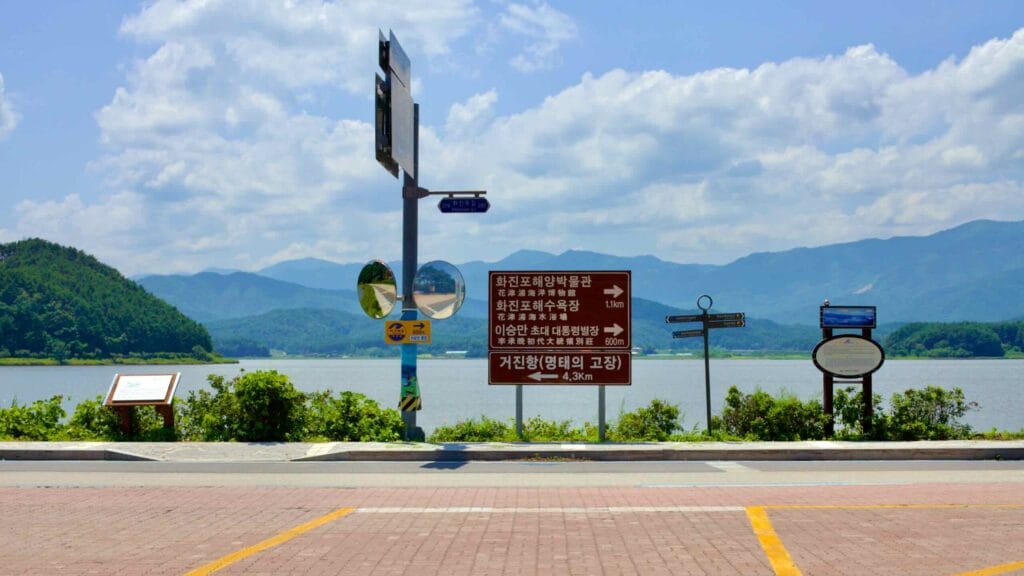A roadside sign in front of Hwajinpo Lake guides visitors towards Geojin Port, Hwajinpo Marine Museum, and Hwajinpo Beach.