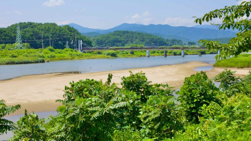 Gazing upriver, the Bukcheon Railroad Bridge emerges in the distance, spanning a stream with prominent sandbanks, all set against a backdrop of green mountains.