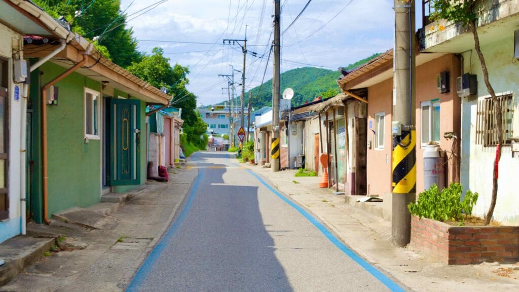 A blue-lined bike path meanders through the heart of Mukho Port, flanked by low houses of the fishing community, modest dwellings, electrical poles, and overhead wires.