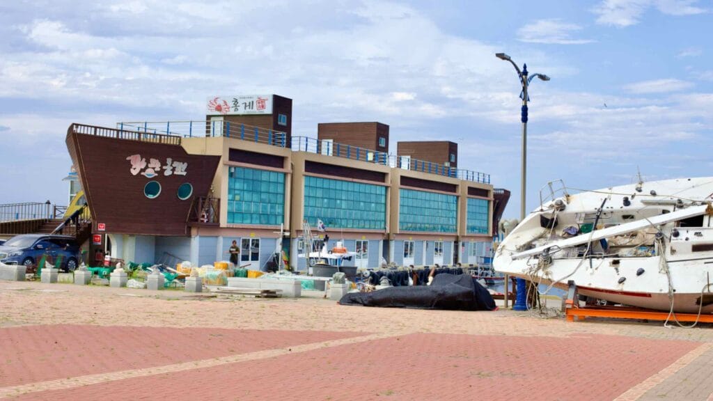 The Daejin Raw Fish Center, a building resembling a large ship and symbolizing Daejin Port, with a sailing boat dry-docked in the courtyard under a clear blue sky.