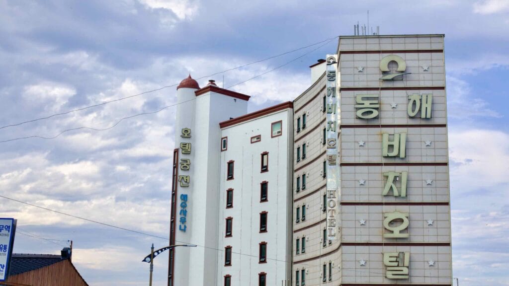 The Donghae Beach Hotel, an old-style tall hotel, stands prominently near Mukho Port under a blue sky with scattered clouds.