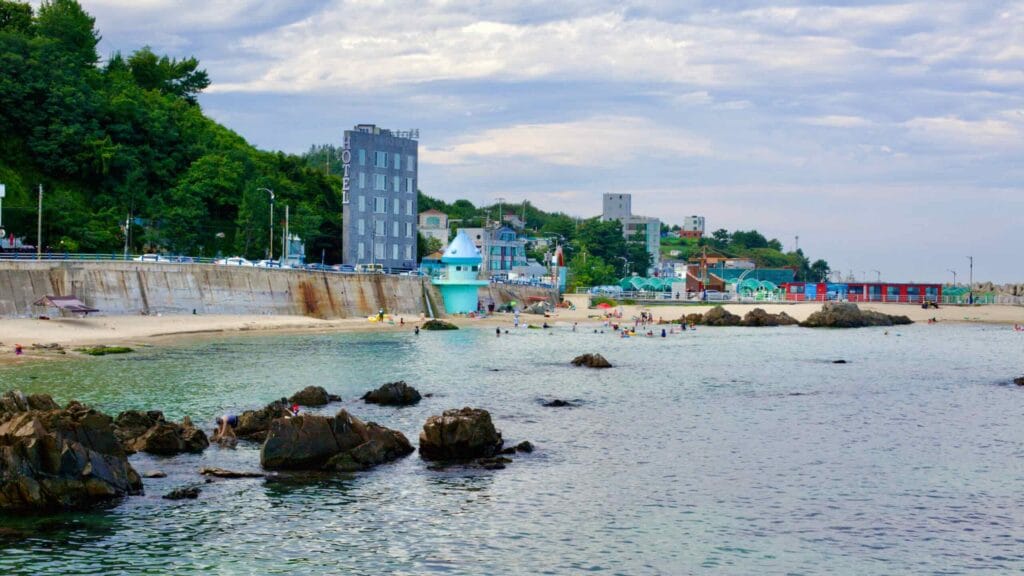 The vibrant scene at Eodal Port and Beach showcases rocks dotting the waters off the sandy shores, with restaurants and small hotels lining the waterfront on a sunny summer day.