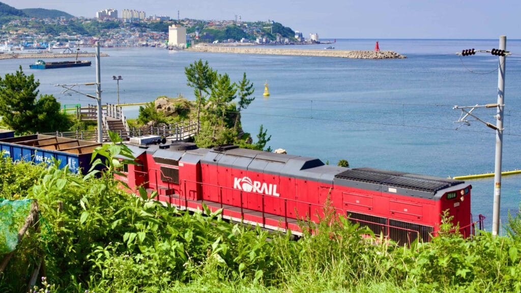 A vibrant red Korail train sweeps past the observation platform on a lush green hillside, offering a dynamic view of Mukho Port, its calm waters, and the distant Mukho Lighthouse.