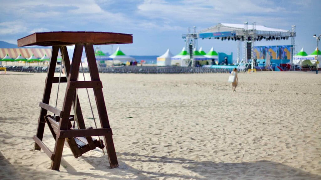 A serene setting with a free-standing porch swing and an open-air performance stage on the sandy grounds of Mangsang Beach, under a clear blue sky on a sunny day.