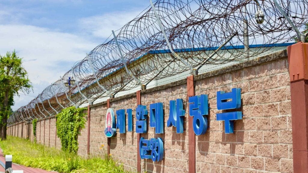 A wall, crowned with razor wire, encircles Donghae Port, under a clear blue sky. A sign mounted on the wall marks the boundary of this secure area.