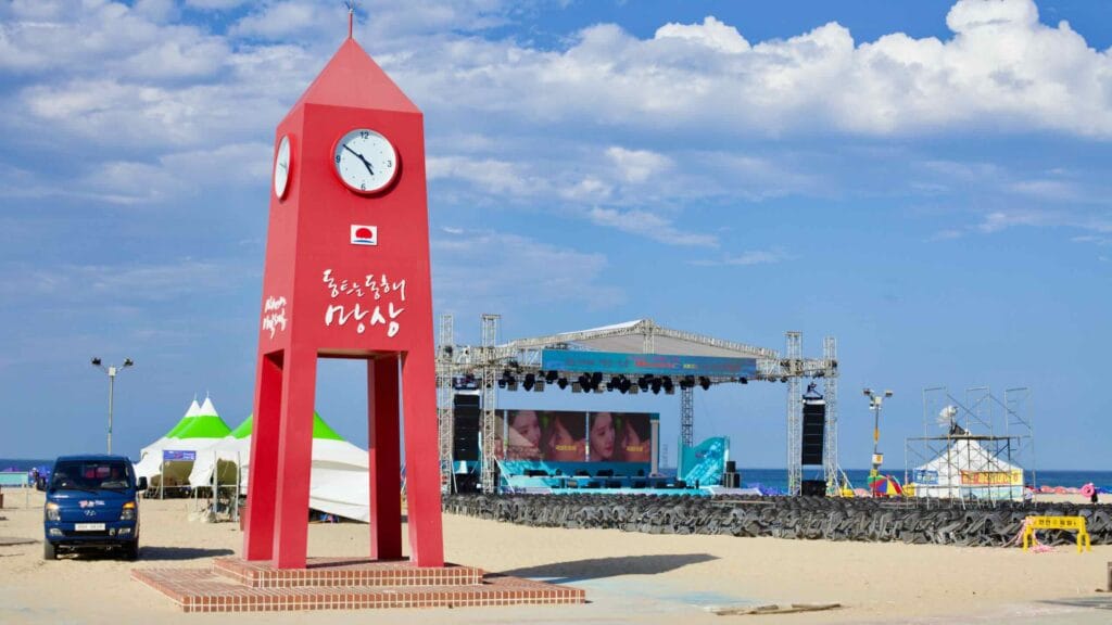 The iconic red clocktower stands tall at Mangsang Beach against a backdrop of an open-air performance stage, under the clear blue sky of a sunny day.