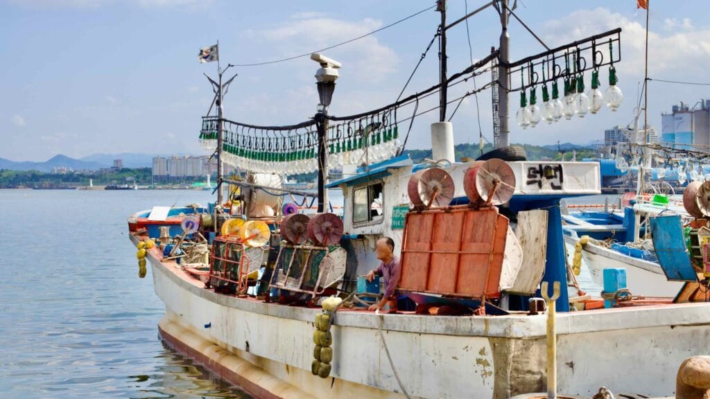 An old, small squid fishing boat moored at Mukho Port, with large lightbulbs strung above the deck to attract squid at night. A fisherman prepares the vessel under the summer sky.