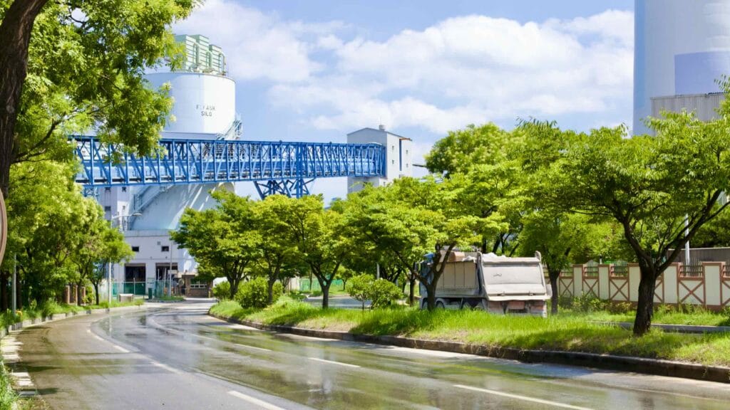 A curving street slices through the industrial heart of Donghae City, within the green-tinged Donghae Free Trade Zone. A dump truck navigates the road, passing under the blue frame of a conveyor belt.