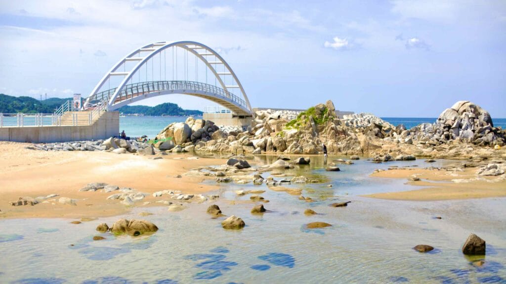 An arched bridge extends to a rocky outcrop off the coast in Namae Port, with transparent waters in the foreground on a summer day.
