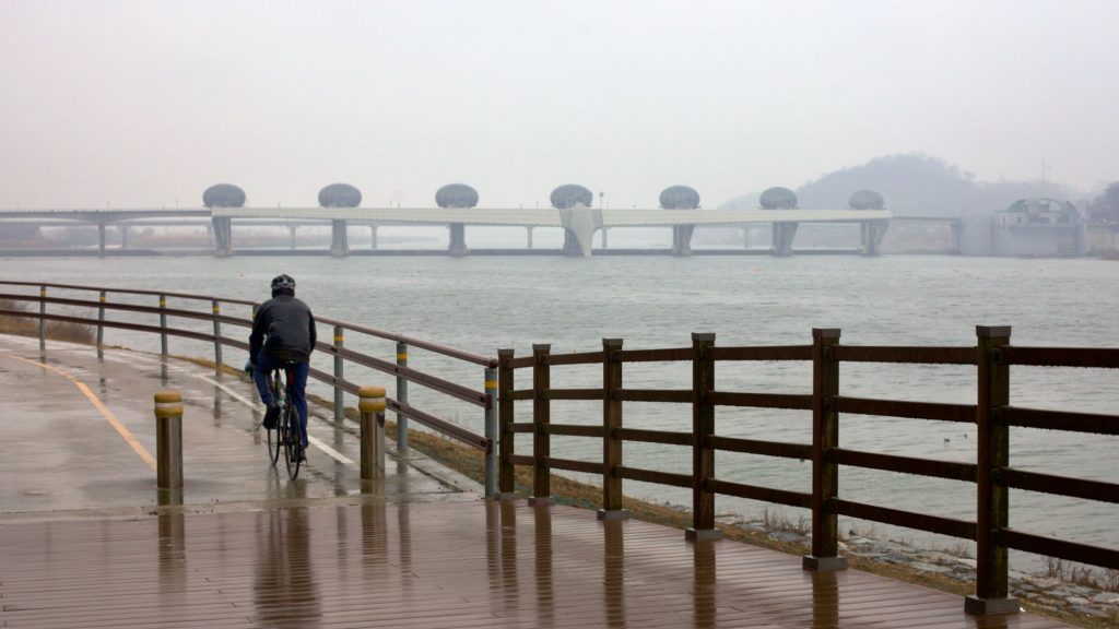 A picture of a cyclist riding through the snowy and rainy day near the Ipo Weir along the Hangang Bicycle Path.