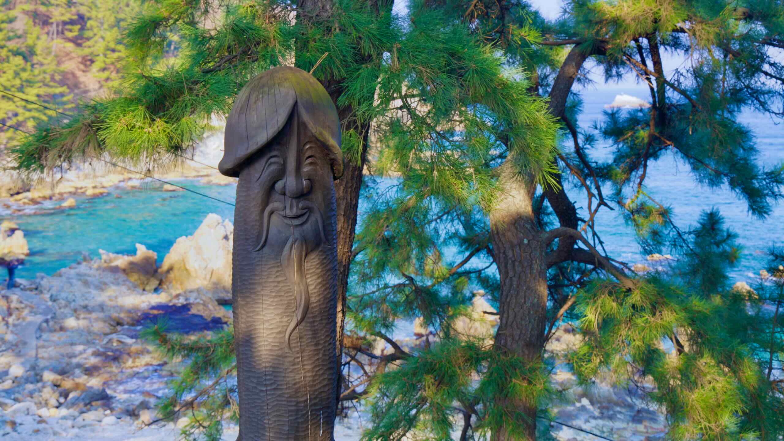 A whimsical sculpture of an old Asian man with a long mustache incorporated into a penis statue, set against a small bay in the East Sea with pine trees and blueish-green waters.