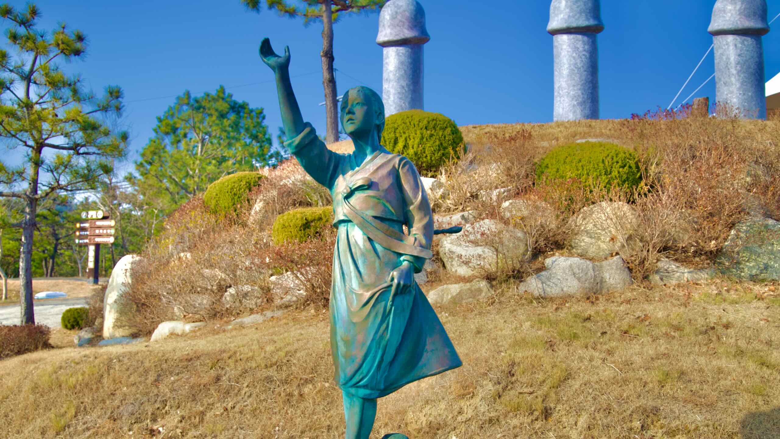 A stone sculpture of the young lost maiden, in the same iconic pose, set against a hillside and surrounded by penis zodiac statues in Haesindang Park.