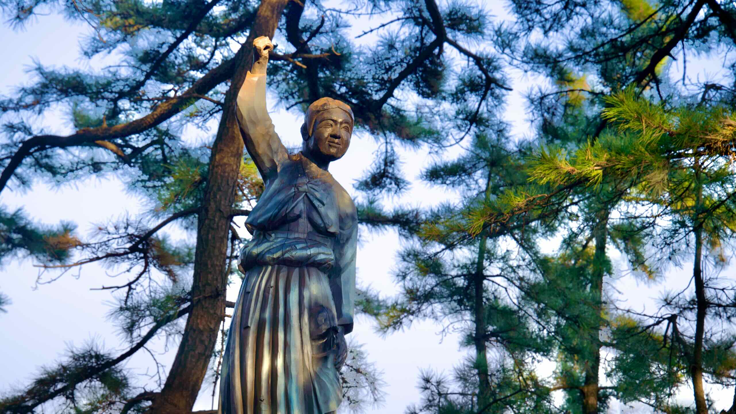 A poignant wooden sculpture of the young lost maiden from local folklore, hand raised, set against a backdrop of pine trees and a blue sky in Haesindang Park.