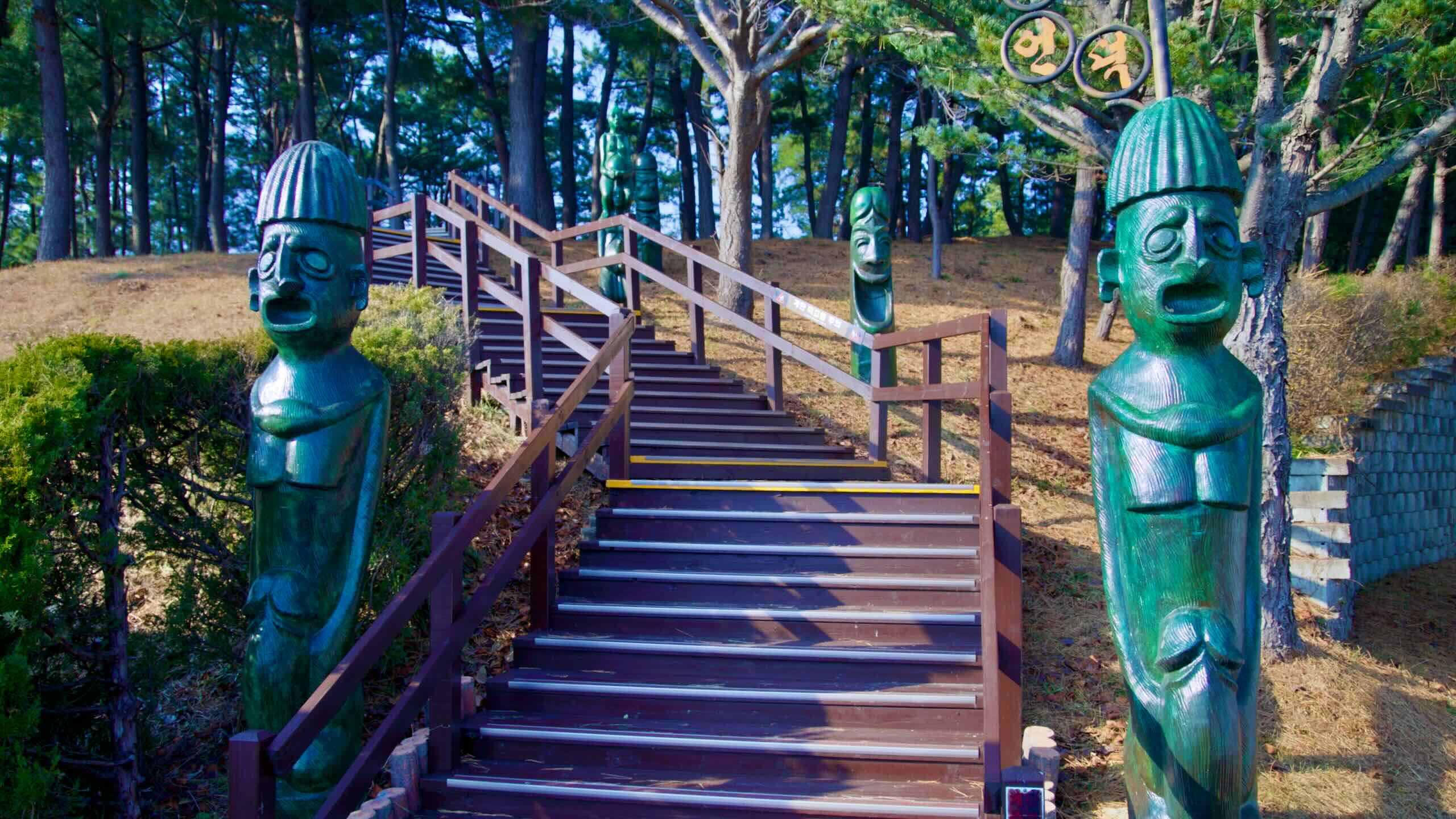 A wooden staircase at Haesindang Park, flanked by distinctive penis statues with human faces, leading up to a serene pine tree forest atop a hill.