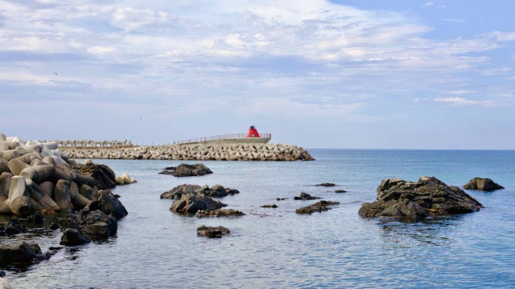 A serene view of the breakwater with low rocks emerging from the East Sea, leading to a quaint red lighthouse at its end.