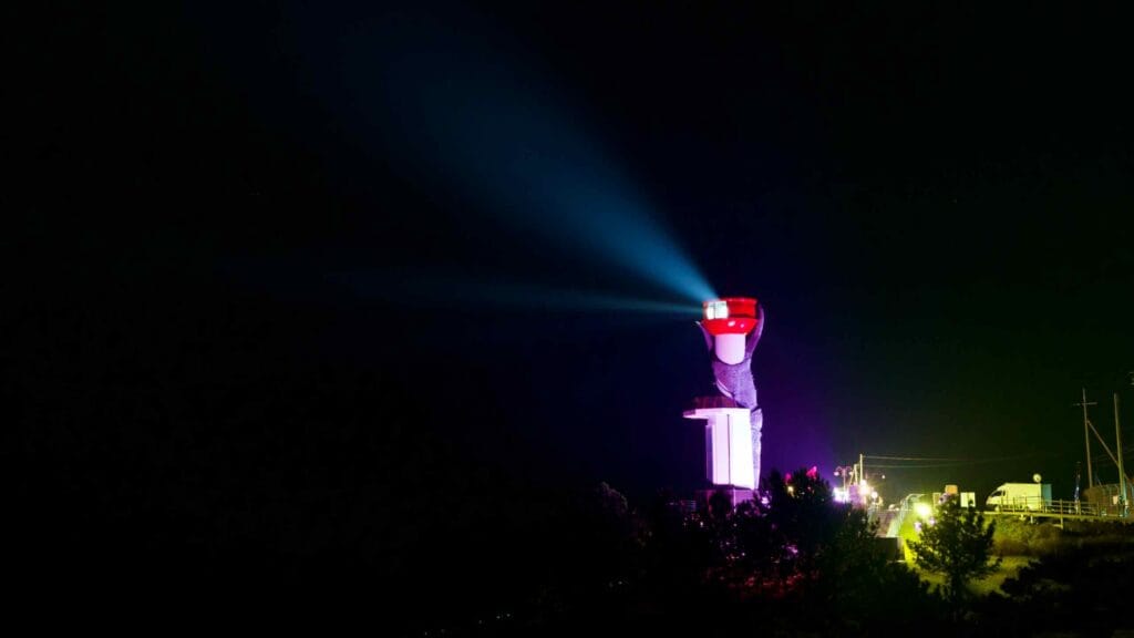 Changpomal Lighthouse stands against the inky black night on a hillside, its light piercing the darkness, with a crab sculpture adorning the structure and cars parked along the roadside below.