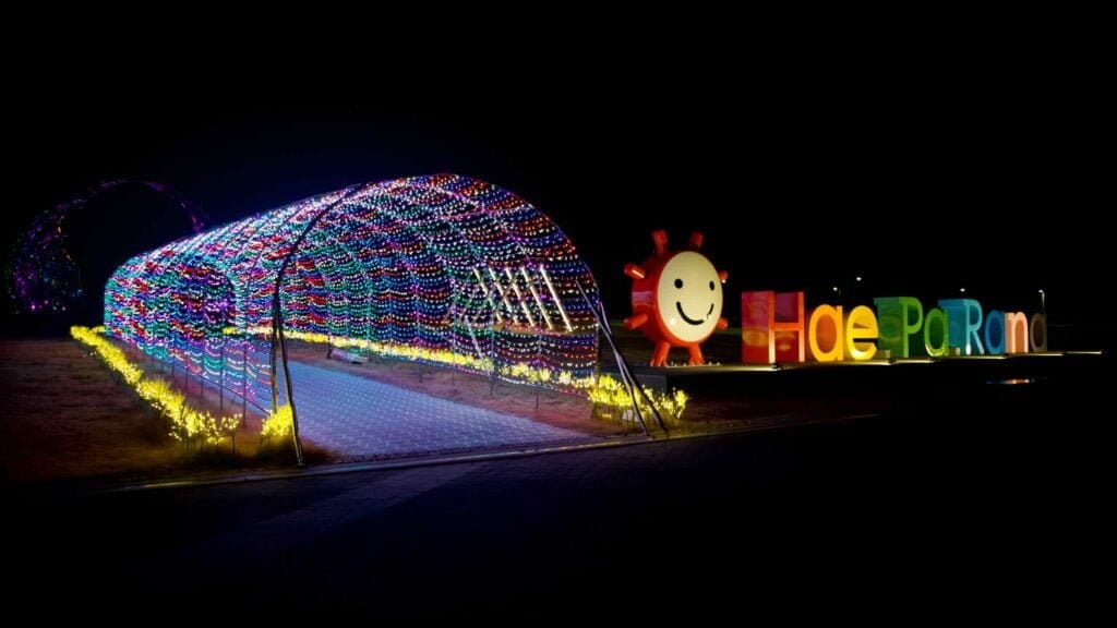 A mesmerizing tunnel of Christmas lights and a vibrant, brightly lit sign captivate visitors in Haeparang Park, adding a festive glow to the seaside ambiance.