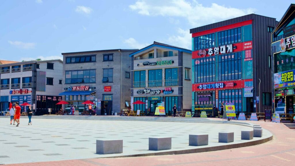 A charming curving courtyard lined with restaurant storefronts at Chuam Beach, featuring two and three-story buildings with glass windows offering panoramic views of the beach.