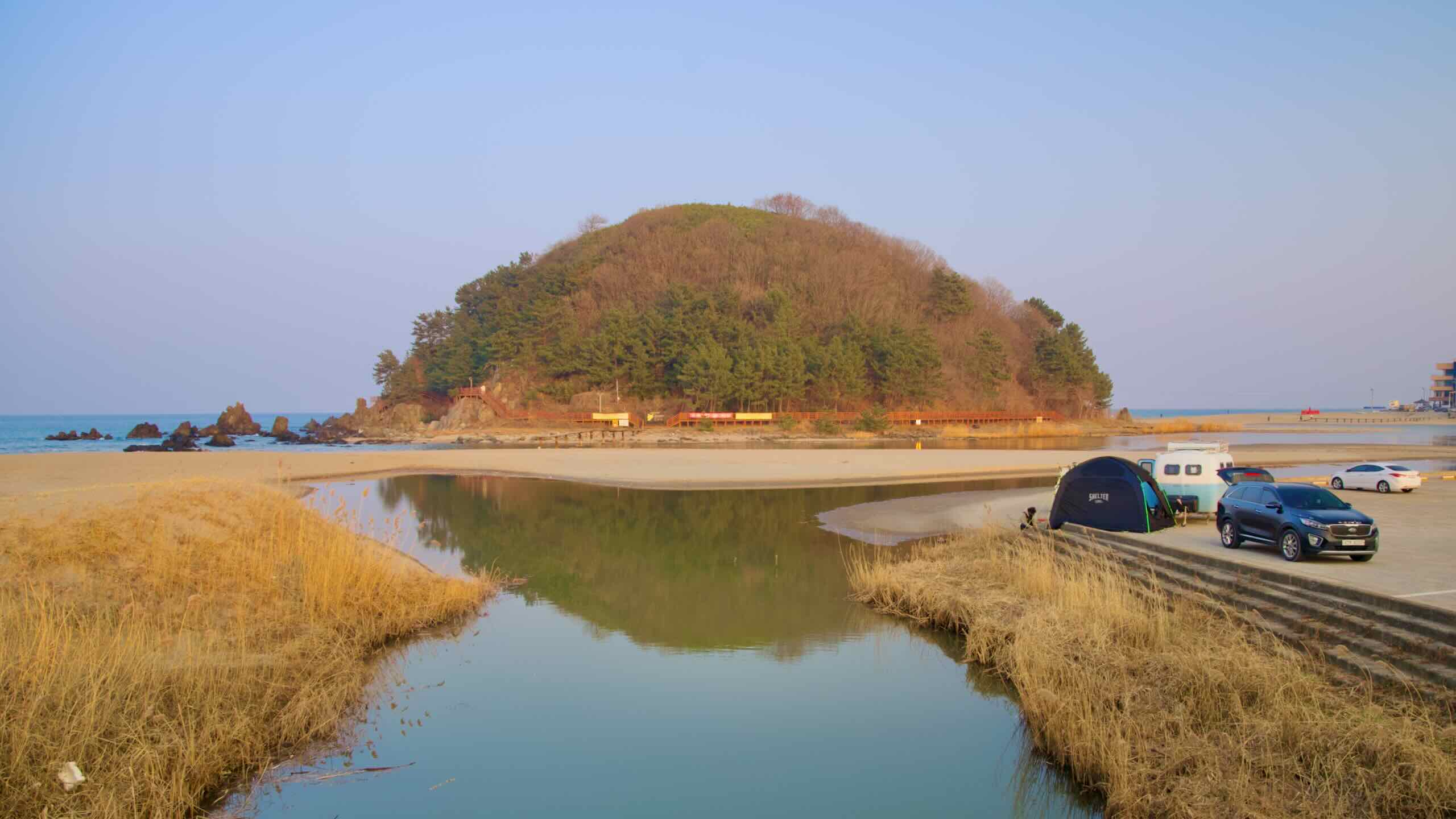 Deokbong Mountain, a round, short hill next to the East Sea, is encircled by a sandy beach with a small stream, and a car with a tent is spotted in the parking lot nearby.
