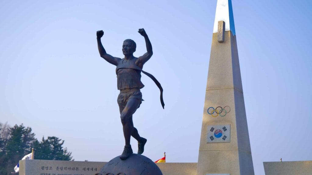 Close-up of Hwang Young-jo statue, depicting him running through finish line tape on a ball with Olympic rings, a tall pyramid, Korean flag, and Olympic rings in the background.
