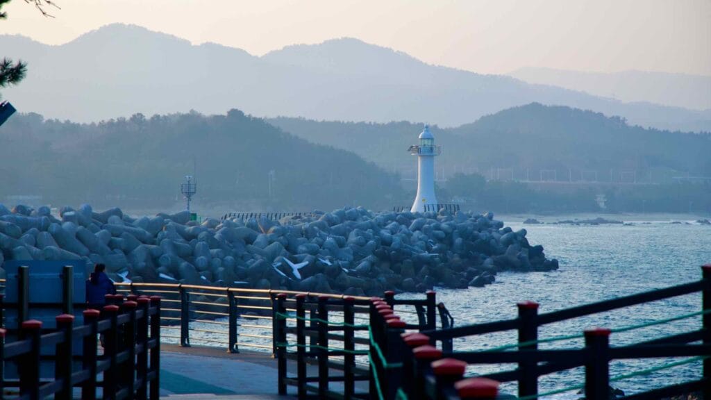The beginning of the Chogok Yonggul Cave Hiking Trail, with the tetrapod breakwater of Chogok Port and its white lighthouse in the background, alongside seaside mountains.