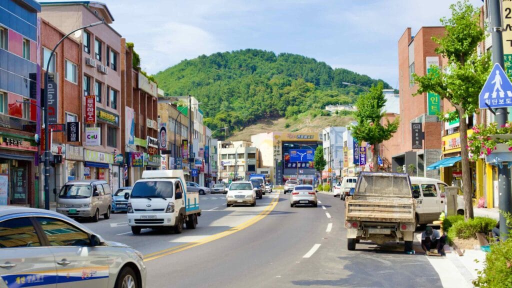The main road cuts through Mukho Port, bordered by three-story stores on either side, with a tree-covered hillside looming in the distance.