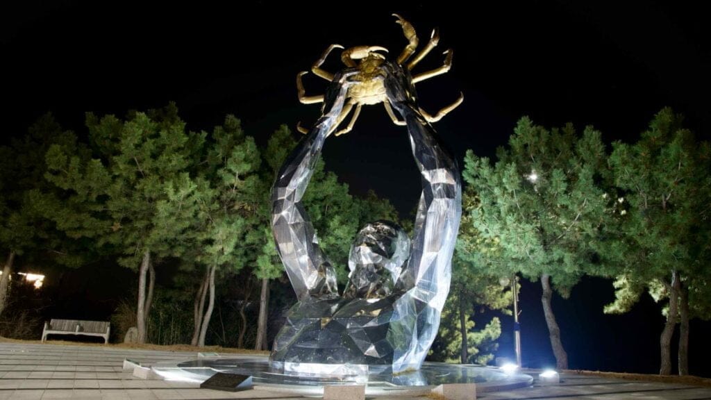 The "Ocean Light" sculpture, depicting a person lifting a giant snow crab, shines under night lights south of Sunrise Park, with trees illuminated in the backdrop.