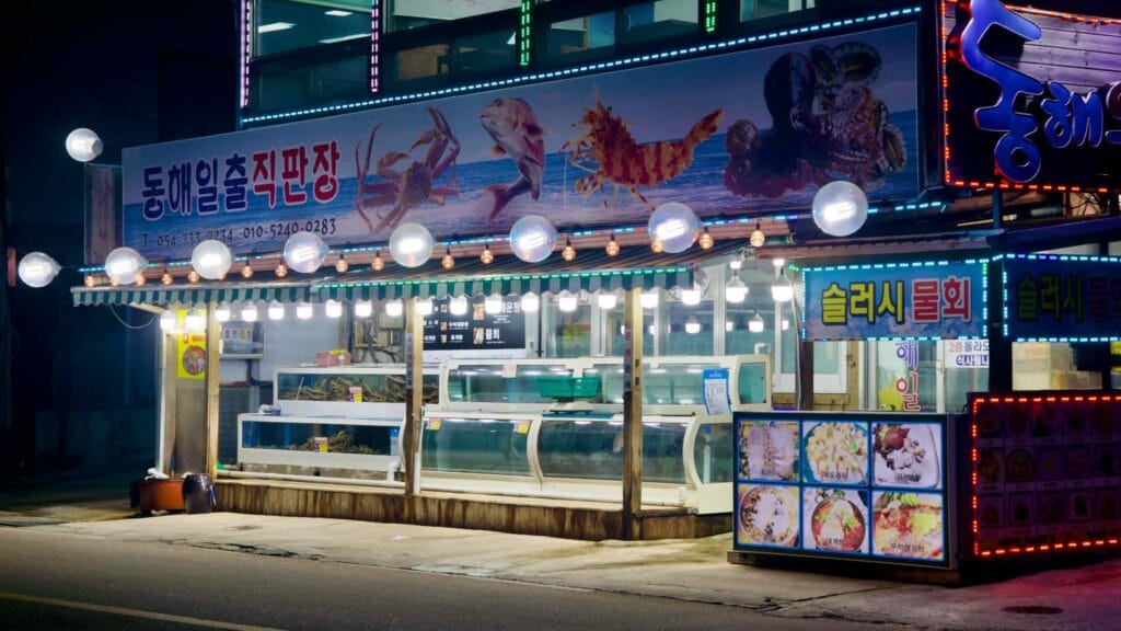 The exterior of a snow crab restaurant at night in Ganggu Port, featuring bright white light bulbs along the awning over water tanks filled with snow crabs.