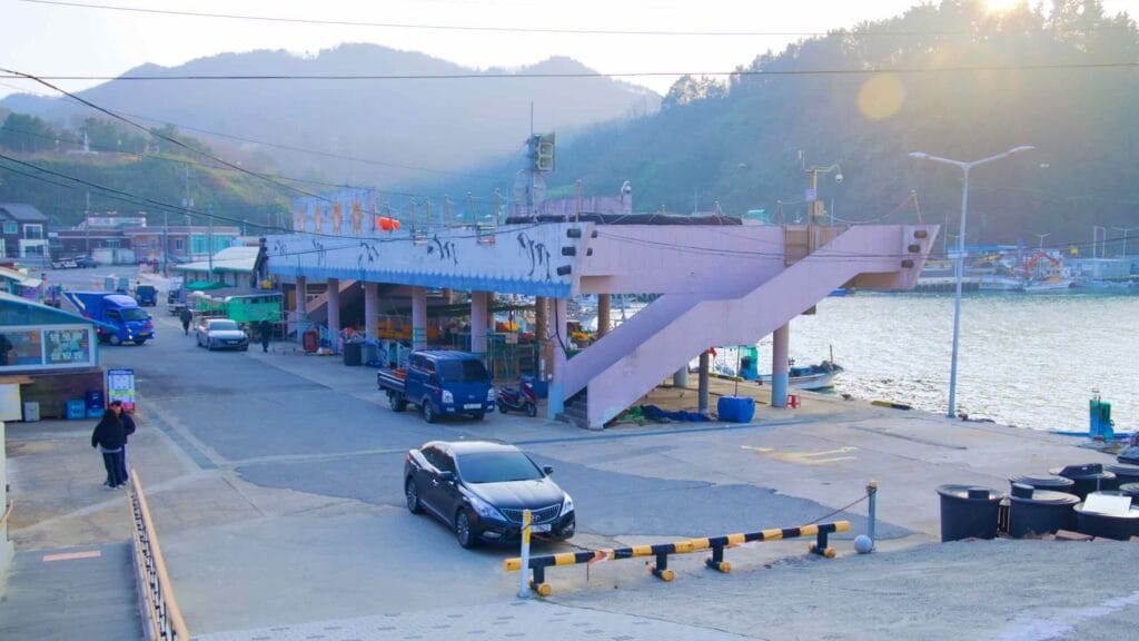 End of day at Chogok Port's seafood market, beside the water with fishing boats nearby, featuring a small open structure and a top deck with drying racks for seafood.
