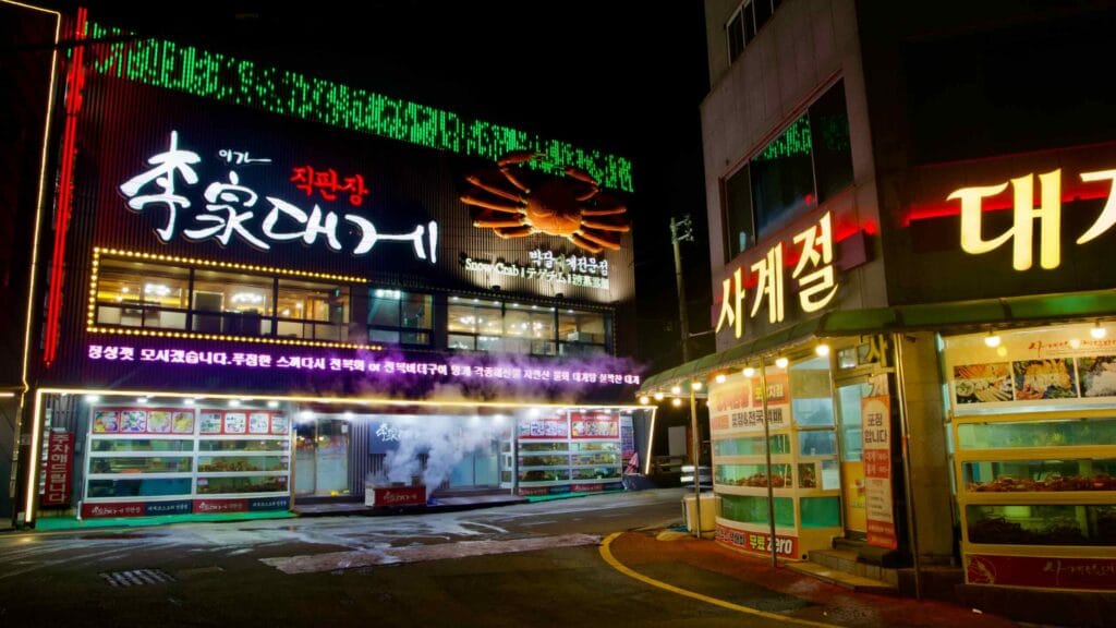 Two snow crab restaurants in Ganggu Port face each other across the street, with steam from crab cookers spewing into the night air and snow crab emblems adorning the sides.