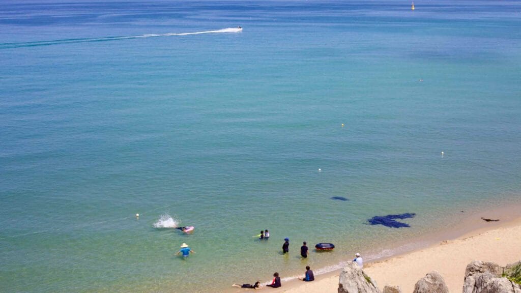 Clear waters at Chuam Beach host swimmers and a distant boat, with a horizon backdrop, rocks on sand, and summer sun.