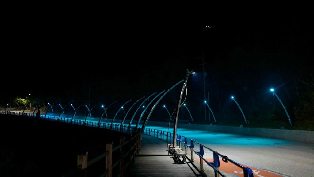 A wide shot of a curving hillside road in Sunrise Park, bathed in turquoise light by LED streetlights, with a walking deck and benches beside the road.