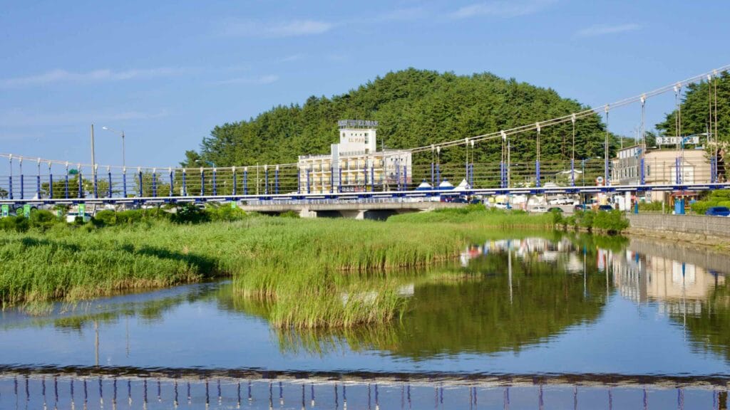 A quaint pedestrian suspension bridge crosses Gwangjeong Stream near Hajodae Beach, with a motel and lush green hill forming a serene backdrop.