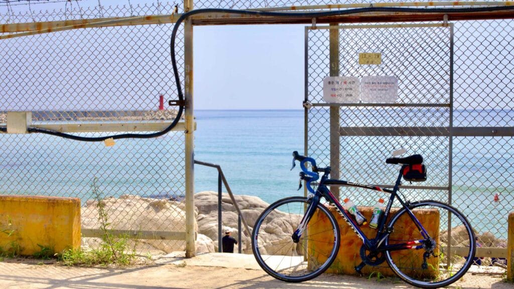 A bike leans against a barbed-wire-topped chainlink fence at Gonghyeonjin Beach, where stairs invite visitors down to a rocky beach below.