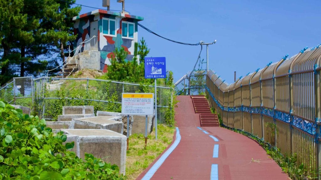 A bike path, delineated by blue lines, guides to a staircase and ramps beside a camouflaged lookout tower near Geojin Village Beach.