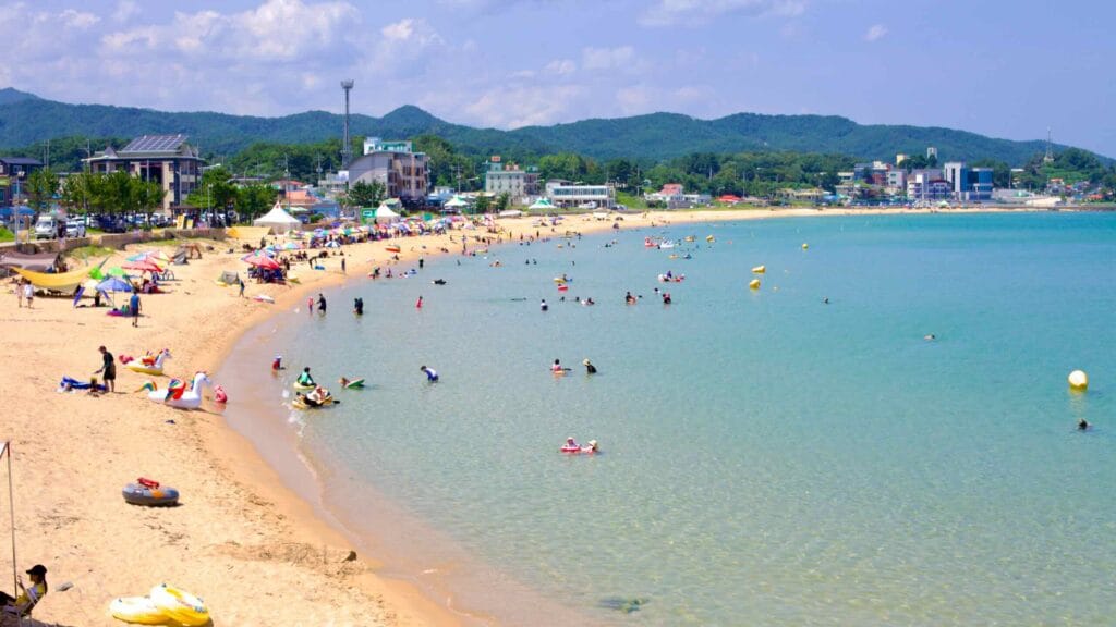Families enjoy the clear waters of Chodo Beach, with its sandy shores framed by seaside hotels and restaurants, and lush green hills rising in the background.