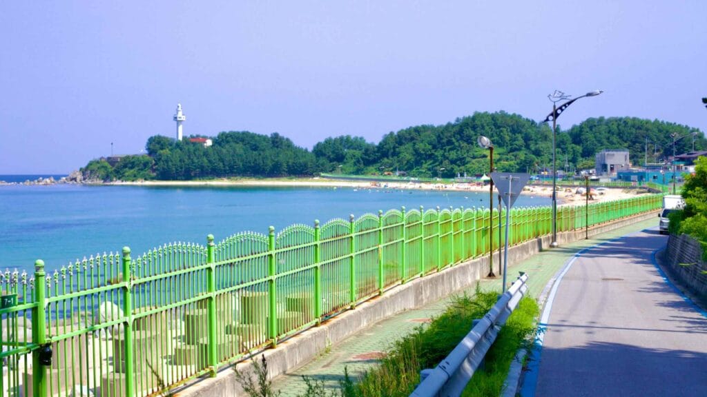 A bike path and road hug the scenic Machajin Beach, bordered by a green fence.