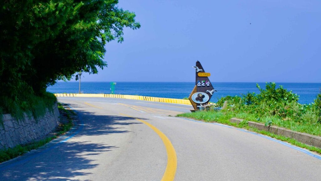 An empty road stretches from Hwajinpo Beach to Chodo Beach, offering breathtaking views of the East Sea under a clear blue sky.