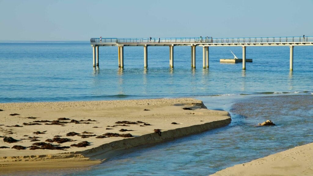 A walkway extends over the serene waters of Jeongdongjin Beach, with a stream cutting through the sandy shore.
